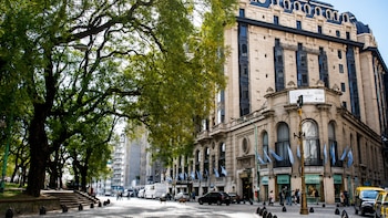 Vista exterior del Plaza Hotel en Buenos Aires. Un edificio neoclásico de piedra con balcones y una cúpula azul. Árboles verdes frondosos y una calle con vehículos y peatones