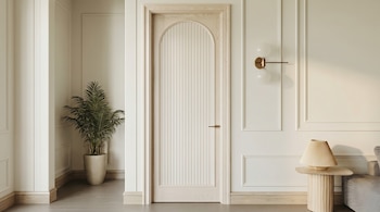 Puerta de madera clara con arco y diseño acanalado. A la izquierda, una planta en maceta. A la derecha, pared con molduras, lámpara y mesa con luz.