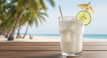 Un vaso de agua de coco cremosa con hielo, una rodaja de lima y sombrilla en una mesa de madera. Fondo borroso de playa y palmeras.