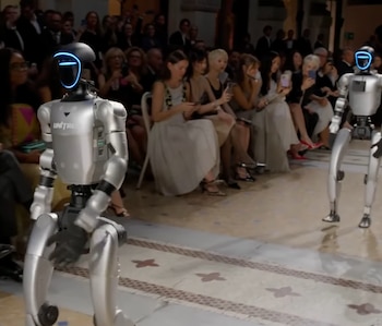 Dos robots metálicos con visor azul iluminado en el rostro caminan por un pasillo entre un público sentado