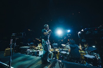Un hombre de pelo gris canta en un micrófono al frente de un escenario oscuro, flanqueado por músicos tocando baterías y percusión bajo potentes luces