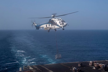 Un helicóptero blanco y azul, modelo Sikorsky S-70, con número de registro N552AC, vuela sobre el mar azul, suspendiendo una jaula de carga con un cable largo