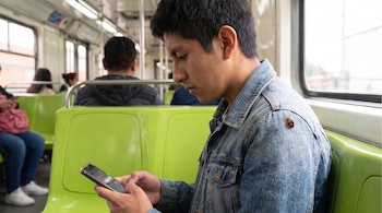 Hombre joven sentado en un vagón de tren, mirando un teléfono móvil, con una chinche pequeña en su hombro derecho, rodeado de asientos verdes.