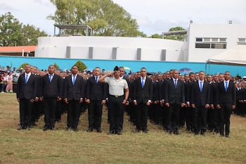 Un grupo de hombres con traje negro y corbata azul, un oficial con uniforme verde y gorra militar, y personas en el fondo sobre césped