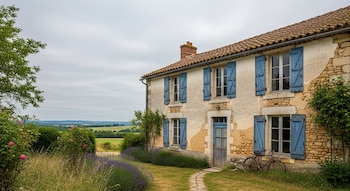 Vista exterior de una casa de campo francesa de piedra con paredes claras y postigos azules en sus ventanas, rodeada de césped, lavanda, rosas y árboles, bajo un cielo nublado.
