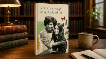 Primer plano de un libro titulado 'Madre Mía' de Mercedes Morán, mostrando a una madre y su hija, con libros, una lámpara y una taza de café en un escritorio de madera