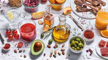 Alimentos como aceite de oliva, aceitunas, gazpacho, carne asada, legumbres, frutos secos y frutas sobre una mesa de madera blanca