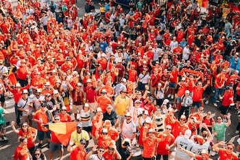 Vista aérea de una gran multitud de personas, la mayoría con camisetas rojas, algunas con banderas belgas, levantando las manos al aire