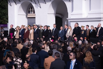 Grupo de funcionarios de gobierno, militares y clérigos de pie frente a un edificio blanco con arcos, observados por una multitud desde abajo