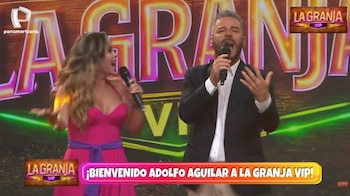 Adolfo Aguilar y una mujer, ambos con micrófonos, cantan en un escenario brillante con el logo "LA GRANJA VIP" y un cartel de bienvenida