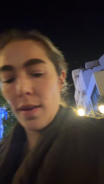 Primer plano de una mujer en una calle de noche, con farolas encendidas y la fachada de un edificio al fondo