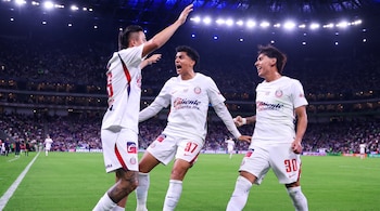 Tres jugadores de fútbol con uniforme blanco gritan y sonríen en una cancha de césped frente a las tribunas iluminadas de un estadio.