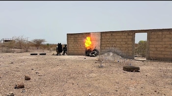 Dos personas con uniforme militar observan fuego cerca de un muro de ladrillos con neumáticos en un terreno árido