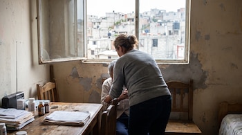 Una mujer de espaldas atiende a una persona sentada en un hogar humilde, con paredes desgastadas y una ventana abierta a una barriada. Documentos y medicinas en la mesa.