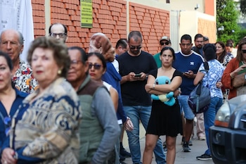 Grupo diverso de personas de varias edades hace fila en exteriores, algunos mirando sus teléfonos, frente a una pared de ladrillo y un vehículo