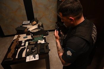 Un policía de espaldas sostiene un radio frente a una mesa con un fusil, un revólver en su estuche, cargadores y cajas de proyectiles