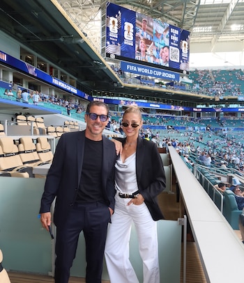 Un hombre y una mujer posan en un estadio con asientos y una pantalla gigante que muestra el logo de FIFA World Cup 2026 y Miami