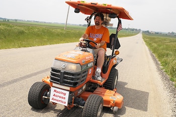 Un hombre con camiseta naranja maneja una cortadora de césped naranja con un techo protector sobre una carretera entre campos