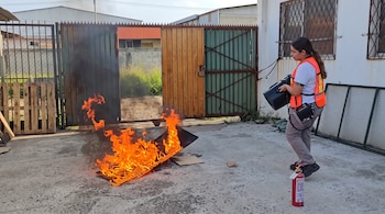Una mujer con chaleco de seguridad y equipo técnico observa cartón ardiendo en el suelo cerca de un extintor en un patio.