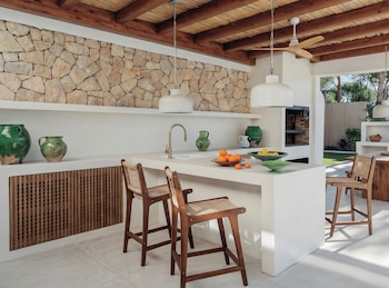 Cozinha exterior com ilha central, três cadeiras em madeira, freixo, parede em pedra, teto em madeira, luminária, candeeiro, grelhador, forno a lenha, mesa de jantar em madeira, bancada e jardim.