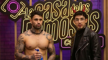 Dos hombres, Tebi Bernal sin camiseta y tatuado, y Valentino Lázaro con chaqueta de cuero y cuernos rojos, posan ante el logo de neón de 'La casa de los famosos Colombia'.