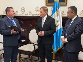 Tres hombres con traje y corbata parados en una sala formal; el de la izquierda sostiene un documento oscuro. Detrás, una bandera de Guatemala