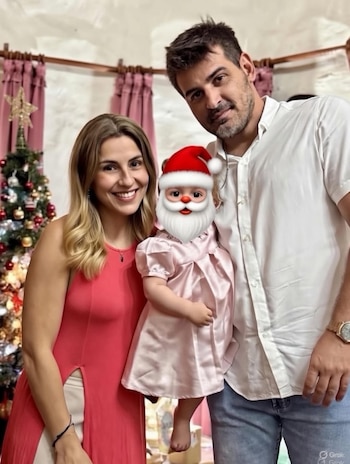 Una mujer y un hombre sonríen a la cámara, sosteniendo a una niña pequeña con un filtro de Papá Noel en la cara, delante de un árbol de Navidad y cortinas rosas