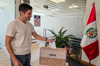 Hombre joven de camiseta blanca sonríe mientras deposita una papeleta en una urna de cartón con el logo de ONPE, con una bandera peruana a la derecha