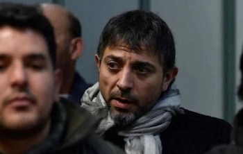 Retrato de primer plano de un hombre de cabello oscuro, barba corta y bufanda clara, rodeado por otras personas en segundo plano