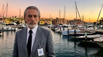 Hombre con traje y corbata posa delante de un puerto deportivo con muchos barcos, edificios coloridos y palmeras al atardecer, bajo un cielo naranja y rosa.