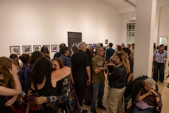 Interior de sala luminosa con paredes blancas y suelo de baldosas claras, mesa de madera, sillas modernas, cuadros, tapices coloridos y dos sillas negras ornamentadas