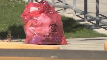 Una bolsa plástica roja con el símbolo de riesgo biológico permanece sobre el suelo junto a una barandilla metálica y césped
