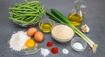 Ingredientes frescos sobre mesada gris: chauchas, huevos, harina, pan rallado, aceites, sal, pimentón, ají molido, cebollas de verdeo y ajo en recipientes.