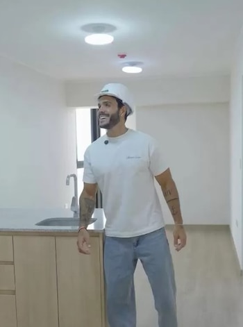 Un hombre con casco blanco de seguridad, camiseta blanca y pantalón de mezclilla sonríe en la cocina de un departamento vacío