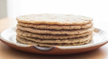 Una pila de siete tortillas de avena de color beige claro apiladas sobre un plato artesanal de cerámica marrón y blanco, con un fondo blanco brillante.