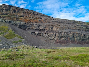 Ladera de una montaña rocosa con estratos sedimentarios visibles y vegetación baja sobre un suelo con pasto bajo un cielo azul