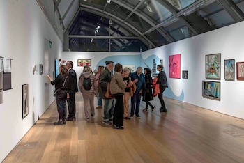 Un grupo de personas camina y observa diversas obras de arte colgadas en paredes blancas de una galería, con un techo de cristal y suelo de madera clara