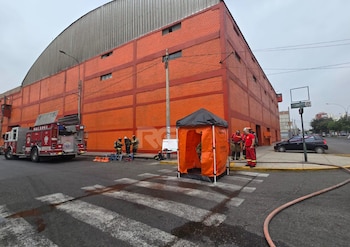 Bomberos y un camión de rescate permanecen junto a una carpa naranja en la calle, frente a un edificio grande de ladrillos rojos
