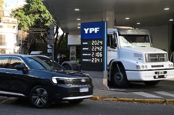 Un SUV Ford oscuro en movimiento pasa un cartel digital de YPF con precios de combustible. Un camión Mercedes-Benz blanco está estacionado en la estación de servicio