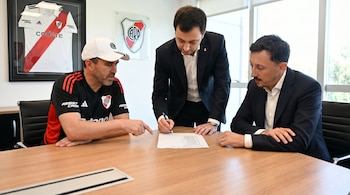 Tres hombres en una mesa de madera. Uno con gorra y camiseta de River Plate señala un papel que otro hombre de traje firma. En la pared, camiseta y escudo de River Plate.