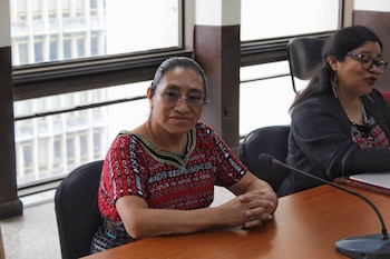 Dos mujeres sentadas ante una mesa de madera con micrófonos; la mujer en primer plano viste una blusa roja bordada y lleva gafas