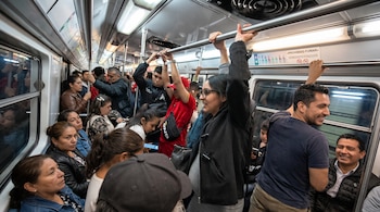 Interior de un vagón de metro con personas sentadas y de pie que se sujetan a las barras metálicas superiores.
