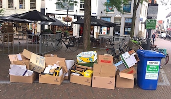 Gran cantidad de cajas de cartón apiladas y otros desechos junto a un contenedor azul de reciclaje en una acera con cafeterías y bicicletas en el fondo