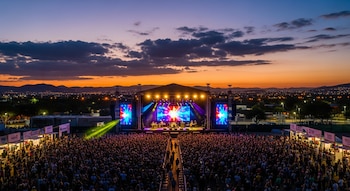 Vista aérea de un concierto al aire libre con un escenario brillante, pantallas LED y una gran multitud de personas, al atardecer con luces de la ciudad de fondo.