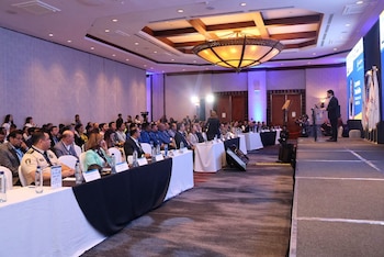 Un expositor junto a un atril y dos pantallas grandes presenta información ante un público sentado en un salón de conferencias