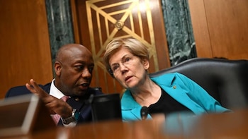 Primer plano de un hombre calvo afroamericano en traje gesticulando mientras habla con una mujer blanca de cabello rubio y gafas con un blazer turquesa