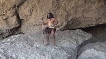 Hombre con cabello largo y semidesnudo, vestido con pieles y sujetando un bastón de madera con un cráneo, en un entorno rocoso