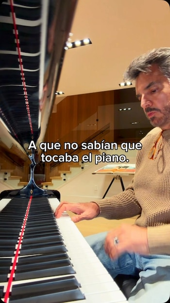 Hombre con barba y cabello canoso toca el teclado de un piano negro, con texto sobreimpreso y una escalera de madera al fondo