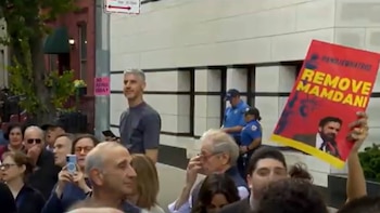 Multitud de personas en una calle con edificios al fondo, con un hombre de cabello gris y un manifestante sosteniendo un cartel rojo que dice "REMOVE MAMDANI"