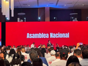 Una multitud asiste a un evento con un orador en el podio frente a una gran pantalla roja que muestra el texto "Asamblea Nacional"